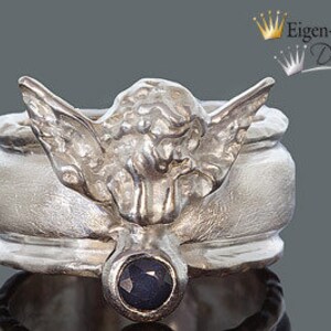 May include: A silver-toned ring featuring a sculpted winged figure with textured details and a single, round, dark blue faceted gemstone set at the base. The band has a matte finish and the text Eigen-Schmied Design is visible in the upper right corner.