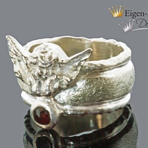 May include: A silver-toned ring featuring a sculpted angel figure with outstretched wings on the band. Below the angel, a small, round, deep red gemstone is set into the metal. The ring has a textured, uneven surface finish throughout.