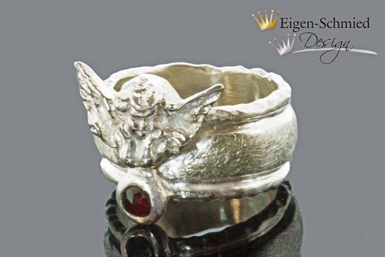 May include: A silver-toned ring featuring a sculpted angel figure with outstretched wings on the band. Below the angel, a small, round, deep red gemstone is set into the metal. The ring has a textured, uneven surface finish throughout.