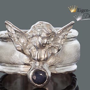 May include: A silver ring featuring a sculpted angel figure with outstretched wings and a single round, dark blue faceted gemstone set at the base. The metal has a brushed, matte finish. The text Eigen-Schmied Design appears in the upper right corner.