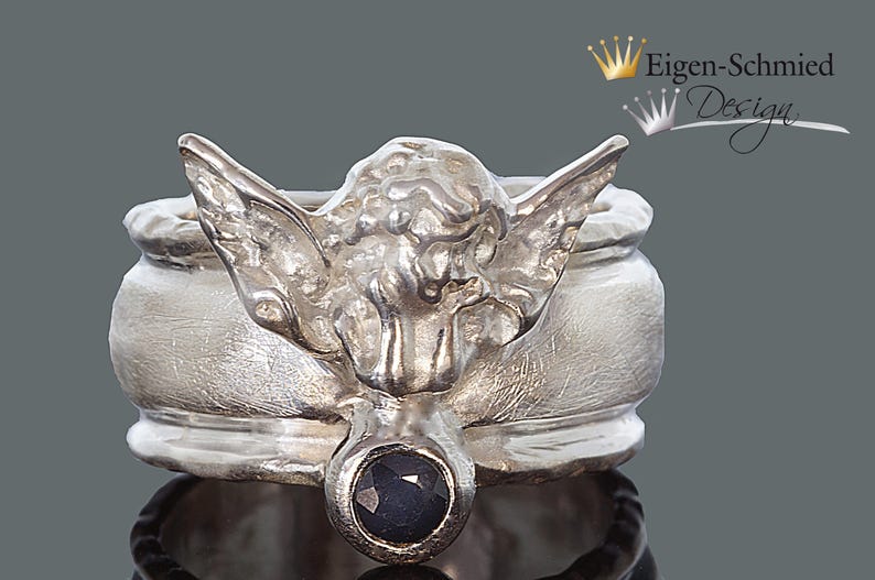 May include: A silver ring featuring a sculpted angel figure with outstretched wings and a single round, dark blue faceted gemstone set at the base. The metal has a brushed, matte finish. The text Eigen-Schmied Design appears in the upper right corner.