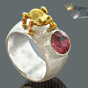 May include: A textured silver ring featuring a small gold-toned frog figure perched on top and a round, faceted dark pink gemstone set into the side. The text Eigen-Schmied Design with two crown icons is visible in the upper right corner of the image.
