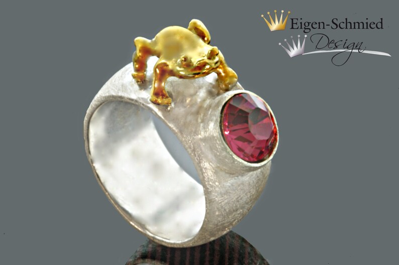 May include: A textured silver ring featuring a small gold-toned frog figure perched on top and a round, faceted dark pink gemstone set into the side. The text Eigen-Schmied Design with two crown icons is visible in the upper right corner of the image.