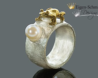 May include: A silver ring with a textured surface, a gold frog on top, and a pearl-like stone on the side. The ring is made by Eigen-Schmied Design.