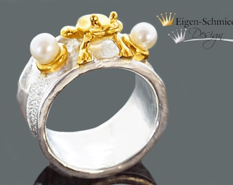 May include: A silver ring with a gold frog and two pearls. The ring is textured and has a wide band. The frog is sitting on top of the ring and the pearls are on either side of the frog. The ring is made by Eigen-Schmied Design.