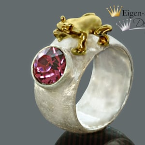 May include: A silver ring with a textured matte finish featuring a round, faceted pink gemstone in a bezel setting and a small, polished gold-colored frog figure perched on the top edge. The text Eigen-Schmied Design is visible in the corner.