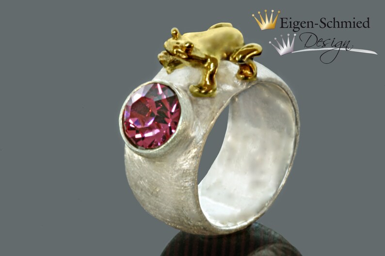 May include: A silver ring with a textured matte finish featuring a round, faceted pink gemstone in a bezel setting and a small, polished gold-colored frog figure perched on the top edge. The text Eigen-Schmied Design is visible in the corner.