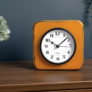May include: A square, orange-yellow clock with a black and white clock face. The clock has a black bezel and white numbers. The clock is on a wooden surface, with a white vase and plant to the left.