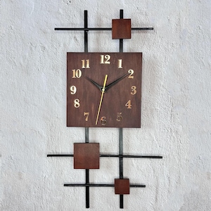 Könnte beinhalten: Eine moderne Wanduhr mit einem dunkelbraunen quadratischen Zifferblatt und goldenen Ziffern. Die Uhr ist von einem schwarzen Metallrahmen mit geometrischen Akzenten umgeben. Die Zeiger sind schwarz mit einem goldenen Sekundenzeiger. Das Design ist minimalistisch und stilvoll.