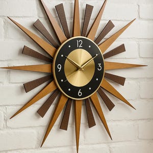 May include: A retro-style wall clock with a sunburst design. The clock has a gold-coloured face with black numerals and hands. The sunburst is made of wood in shades of brown and tan. The clock is mounted on a white brick wall.