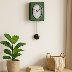 Pode incluir: Um relógio de pêndulo quadrado verde com um mostrador branco e ponteiros pretos pendurado em uma parede branca. O relógio está acima de uma mesa de madeira com uma planta em vaso, uma pilha de livros e uma cesta de vime.