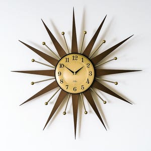 Könnte beinhalten: Eine Retro-Starburst-Wanduhr mit goldenem Zifferblatt und schwarzen Zeigern. Die Uhr hat braune Holzstrahlen und goldfarbene Kugelakzente. Die Uhr ist an einer weißen Wand montiert.