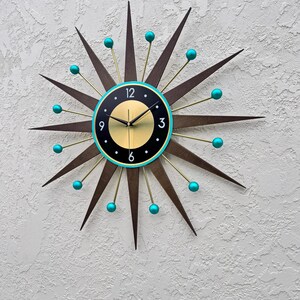 May include: A large, starburst-shaped clock with a black face and gold accents. The clock has turquoise blue accents on the tips of the starburst and around the face. The clock face has black Roman numeral hour markers and gold hands.