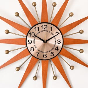 May include: A mid-century modern starburst clock with a copper-colored face and black numerals. The clock features orange wooden rays and gold-colored metal accents. A stylish wall clock for home decor.