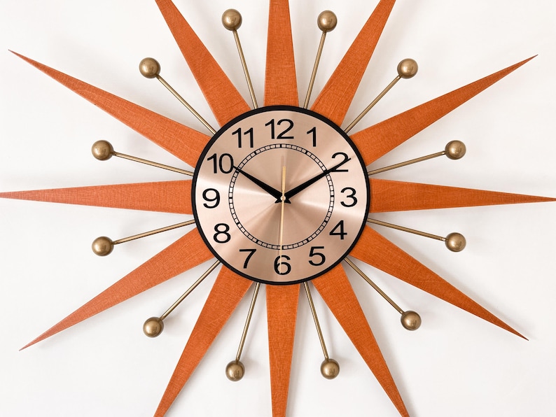 May include: A mid-century modern starburst clock with a copper-colored face and black numerals. The clock features orange wooden rays and gold-colored metal accents. A stylish wall clock for home decor.