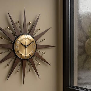 May include: A mid-century modern wall clock with a starburst design. The clock features a gold-colored face with black numerals and hands, surrounded by dark brown wooden spikes and brass accents. The clock is mounted on a beige wall.
