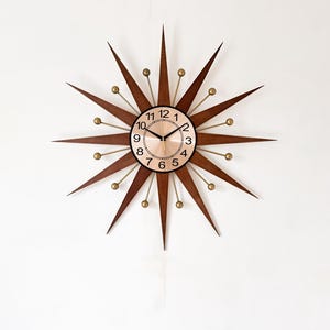 May include: A mid-century modern starburst clock with a brown wooden frame and gold accents. The clock face is a light copper color with black numerals and hands. The clock is mounted on a white wall.