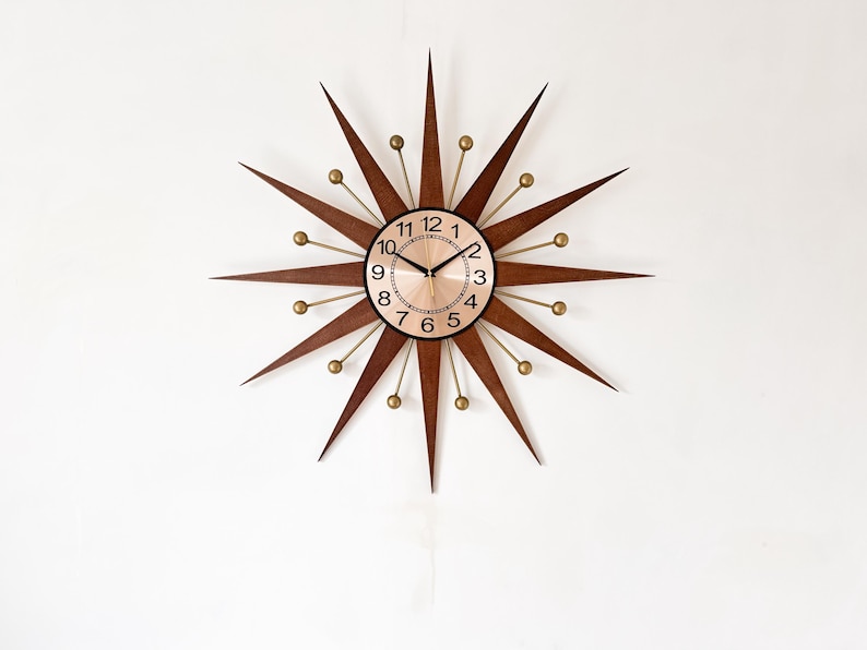 May include: A mid-century modern starburst clock with a brown wooden frame and gold accents. The clock face is a light copper color with black numerals and hands. The clock is mounted on a white wall.