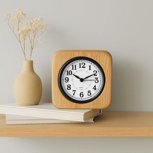 Puede incluir: Un reloj despertador de madera con una esfera blanca y agujas negras. El reloj tiene números negros y un borde negro alrededor de la esfera. El reloj está sentado en un estante de madera.