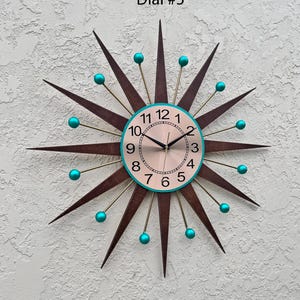 May include: A mid-century modern style wall clock with a starburst design. The clock face is cream colored with black hands and teal blue numbers. The clock has a brown wood frame with gold accents and teal blue balls at the ends of the points.