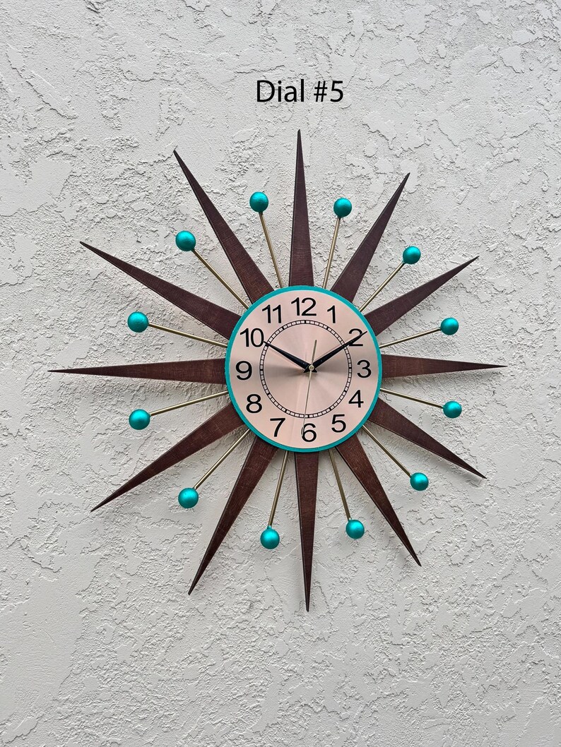 May include: A mid-century modern style wall clock with a starburst design. The clock face is cream colored with black hands and teal blue numbers. The clock has a brown wood frame with gold accents and teal blue balls at the ends of the points.