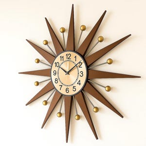 May include: A starburst-shaped wall clock with a light wood-tone face and black hands. The clock features brown wooden rays with gold-colored ball accents. The clock's numbers are black, and the frame is black.