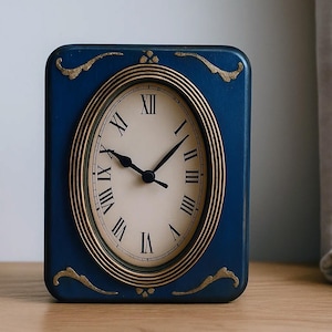Puede incluir: Un reloj rectangular azul marino con una esfera ovalada. La esfera tiene números romanos negros y manecillas negras. El reloj está adornado con dorado y tiene detalles dorados en las esquinas.