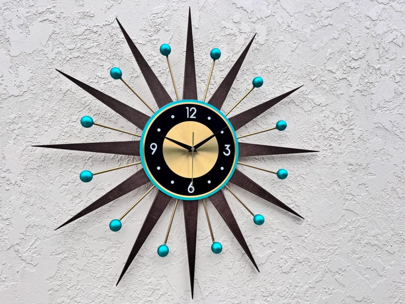 May include: A mid-century modern style wall clock with a black face and gold hands. The clock has a turquoise rim and 12 gold-tipped rays with turquoise balls at the ends.