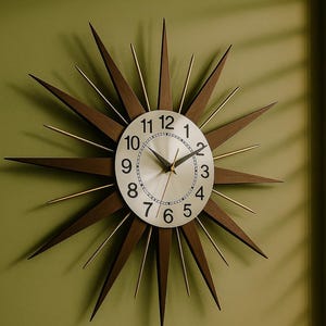 Könnte beinhalten: Eine sternförmige Wanduhr mit silbernem Zifferblatt und schwarzen Ziffern. Die Zeiger der Uhr sind schwarz, und der Rahmen hat dunkelbraune Holzspitzen mit goldfarbenen Akzenten. Die Uhr ist an einer grünen Wand befestigt.
