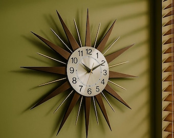 May include: A starburst wall clock with a silver face and black numerals. The clock's hands are black, and the frame features dark brown wooden points with gold-colored accents. The clock is mounted on a green wall.