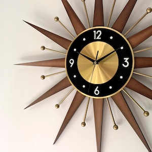 May include: A mid-century modern starburst clock with a gold-tone center and black face. The clock features a gold-tone frame, brown wooden starburst arms, and gold-tone accents. The clock hands are black.