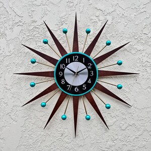 May include: A mid-century modern starburst clock with turquoise accents. The clock face is black with white numbers and a white background. The clock hands are black.