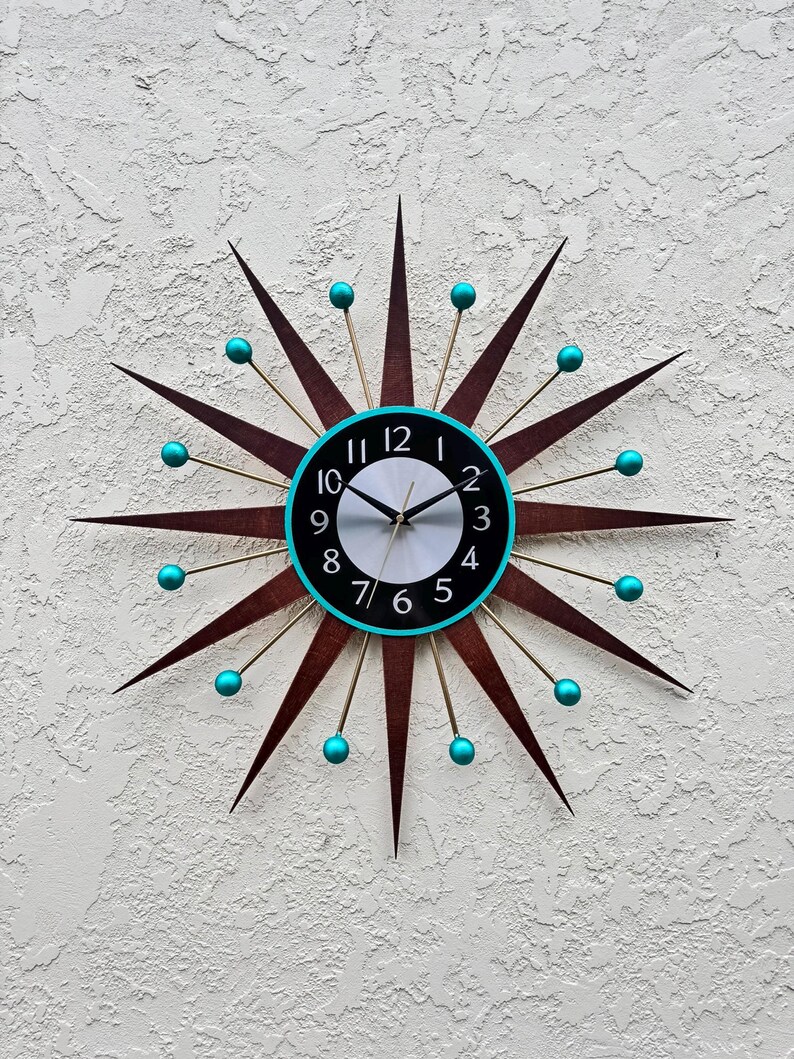 May include: A mid-century modern starburst clock with turquoise accents. The clock face is black with white numbers and a white background. The clock hands are black.