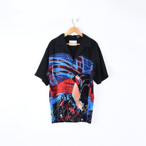 May include: Black short-sleeved button-down shirt with a colorful abstract design. The design features red, blue, and orange shapes. The shirt is hanging on a wooden hanger against a white background.