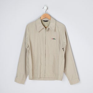 May include: A beige zip-up jacket with a small embroidered logo on the left chest. The logo is green, red, and white.