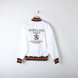 May include: White satin bomber jacket with black and orange striped trim. The back of the jacket features an embroidered logo that says "Maryland Amateur State Jousting Champion".