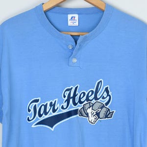 Puede incluir: Camiseta Henley azul claro de manga corta con las palabras "Tar Heels" en cursiva y un gr&aacute;fico de carnero. La camiseta tiene una tapeta de tres botones y cuello redondo. La camiseta est&aacute; hecha de un material suave.