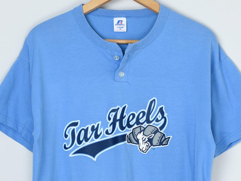 Puede incluir: Camiseta Henley azul claro de manga corta con las palabras "Tar Heels" en cursiva y un gr&aacute;fico de carnero. La camiseta tiene una tapeta de tres botones y cuello redondo. La camiseta est&aacute; hecha de un material suave.