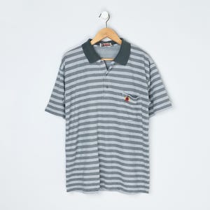 May include: A grey and white striped polo shirt with a dark green collar and a small pocket on the chest. The shirt is hanging on a wooden hanger against a white background. The shirt has short sleeves.