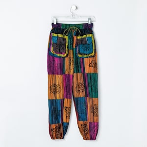May include: Multi-coloured patchwork trousers featuring a drawstring waist, elastic cuffs, and two front pockets. The fabric displays a vibrant striped pattern in shades of orange, purple, blue, and green, accented with black line-art graphic prints.