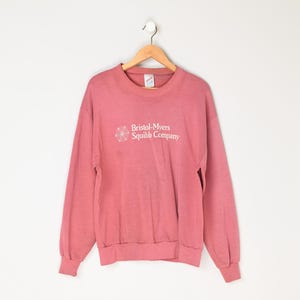 May include: A pink long-sleeve sweatshirt with the text "Bristol-Myers Squibb Company" in white on the front.