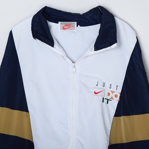 May include: A white, blue, and gold windbreaker jacket with a zipper closure. The jacket has a Nike logo and the text "JUST DO IT" on the front.
