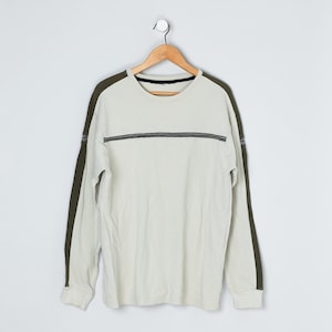May include: A long-sleeve crew neck shirt in off-white with dark olive green panels running down the outer length of each sleeve. A thin, horizontal dark stripe with white stitching detail crosses the chest area of the garment.