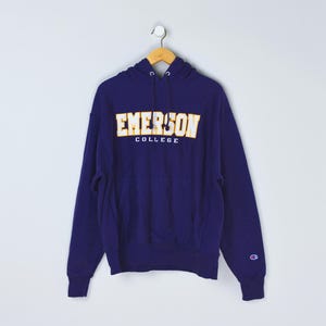 May include: A navy blue hooded sweatshirt with the text "EMERSON COLLEGE" in gold and white lettering. The sweatshirt has a front pocket and a Champion logo on the left sleeve. It is hanging on a wooden hanger.