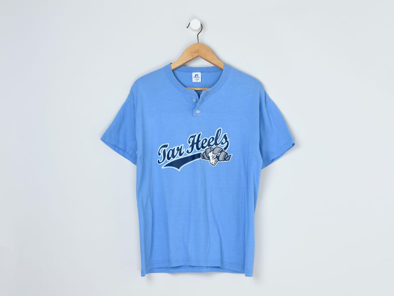 Puede incluir: Camiseta azul claro de manga corta con cuello henley. La camiseta presenta el texto "Tar Heels" en una fuente de escritura con un gr&aacute;fico de carnero. La camiseta est&aacute; colgada de una percha de madera sobre un fondo blanco.