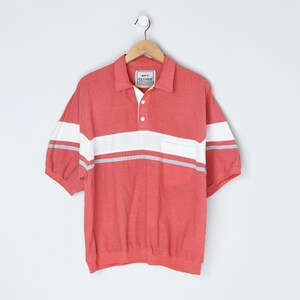 Peut inclure: Un polo de couleur corail avec des rayures horizontales blanches et grises sur la poitrine et les manches. La chemise a un col blanc, une poche poitrine et des manches courtes. Suspendu à un cintre en bois.