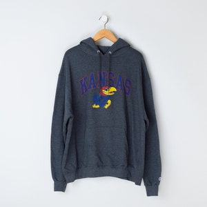 May include: A gray hooded sweatshirt with the word "Kansas" and a cartoon mascot in blue, red, and yellow. The sweatshirt has a pouch pocket and a Champion logo on the bottom right.