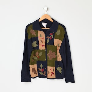 Op de afbeelding: Een marineblauwe cardigan met een patchwork-ontwerp met geborduurde bladeren en bloemen in tinten groen, bruin en rood.