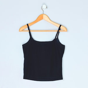 May include: A black camisole top with thin straps hangs on a wooden hanger against a light blue wall. The top is a solid color, with a simple, classic design. The hanger is attached to the wall with a small metal hook.