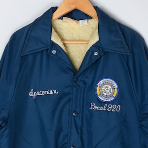May include: A blue jacket with a white fleece lining. The jacket has a white embroidered logo on the left chest that says "Spaceman." The right chest has a yellow and blue embroidered logo that says "Intl Brotherhood of Boilermakers Local 920." The jacket has a Canadian flag patch on the left sleeve.